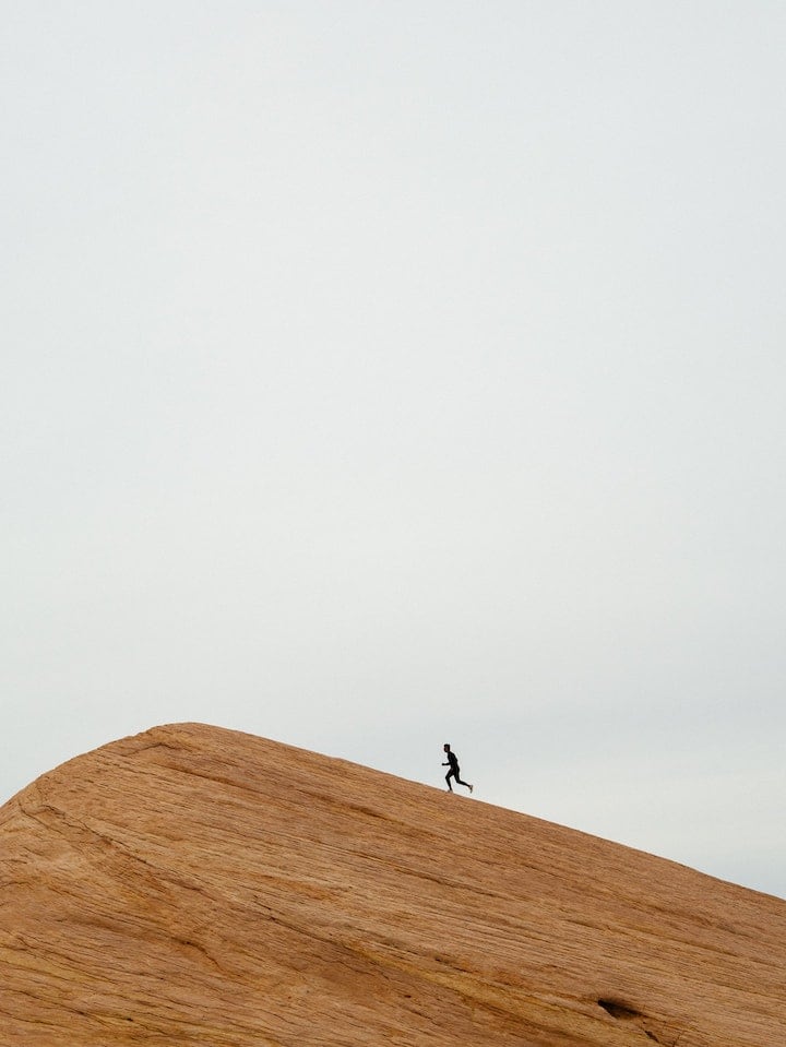 At a distance, solitary man runs up a rock formation, silhouetted against a gray sky.